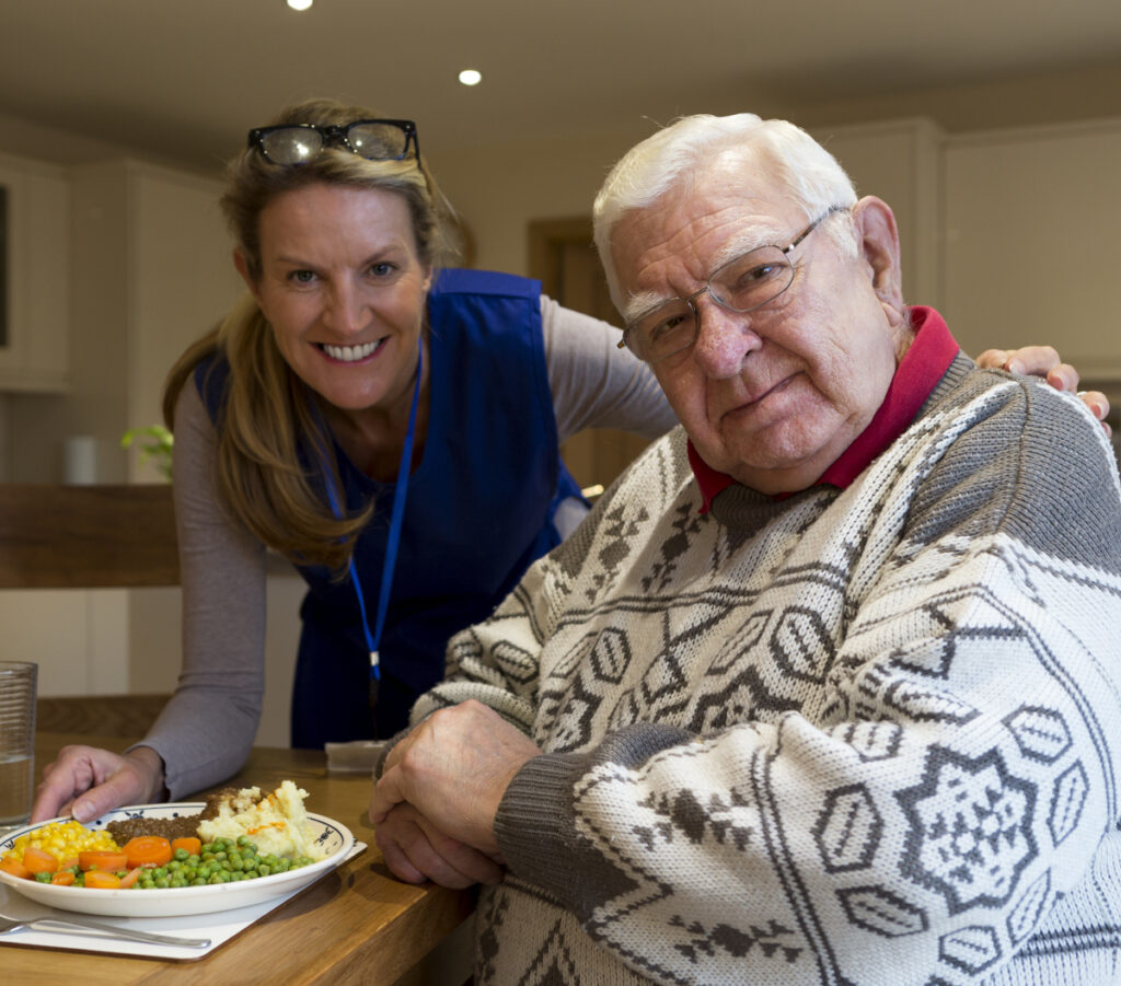 A photograph of a care worker placing a meal in front of an elderly gentleman